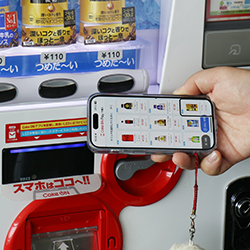 写真：自販機の前で商品一覧の情報をスマートフォンで確認している